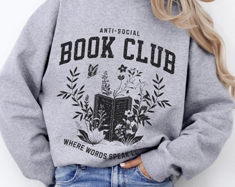 May include: Sport grey sweatshirt featuring the text "ANTI-SOCIAL BOOK CLUB" above a graphic of an open book with floral and butterfly accents. The phrase "WHERE WORDS SPEAK LOUD" is below the graphic. The sweatshirt is worn with blue jeans.