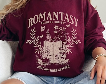 May include: A maroon sweatshirt featuring a graphic design of a book with flowers and butterflies. The text on the sweatshirt reads "Romantasy Readers Society" and "Just One More Chapter".
