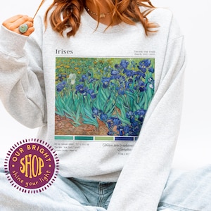 May include: Ash-colored sweatshirt featuring a print of Vincent van Gogh's "Irises" painting. The artwork depicts blue and green irises in a field. The sweatshirt has the text "Irises" and "Our Bright Shop" on it.