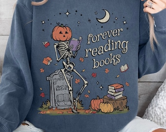 May include: Denim blue sweatshirt with a graphic of a skeleton with a pumpkin head reading a book. The text "forever reading books" is printed on the front. The design includes a tombstone, pumpkins, and autumn leaves.