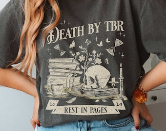 May include: A dark grey t-shirt with a vintage-style graphic of a skull surrounded by books, flowers, and a candle. The text "DEATH BY TBR" and "REST IN PAGES" is printed on the shirt.