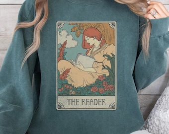 May include: A teal blue sweatshirt with a vintage-style illustration of a woman reading a book in a garden. The illustration is surrounded by a decorative border and the text "THE READER" is printed below it.