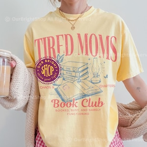 May include: A yellow t-shirt with the text "TIRED MOMS" in red, and "Book Club" in red. The shirt features a graphic of books and a lamp. The word "BANANA" is printed on the bottom right corner.