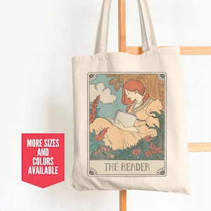 May include: A beige tote bag with a vintage-style illustration of a woman reading a book in a garden. The illustration is surrounded by a decorative border and the text "THE READER" is printed below it.