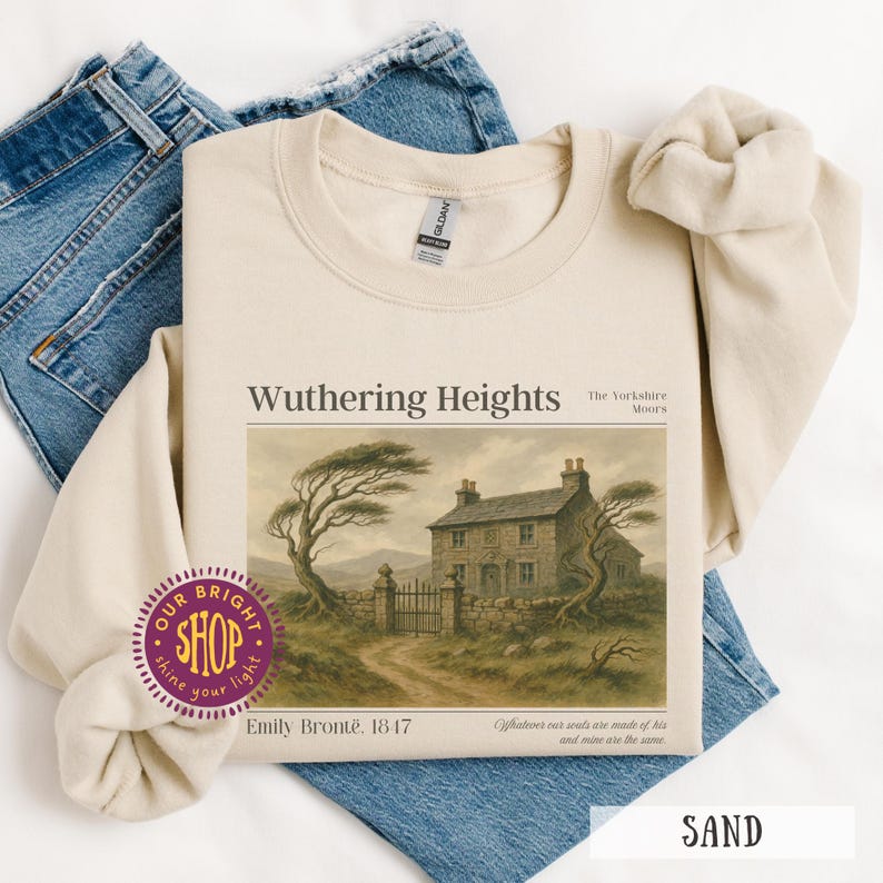Puede incluir: Sudadera color arena con el t&iacute;tulo "Wuthering Heights" y una ilustraci&oacute;n de una casa y un paisaje. Tambi&eacute;n es visible el texto "Emily Bront&euml;, 1847". La sudadera est&aacute; doblada sobre unos vaqueros azules.
