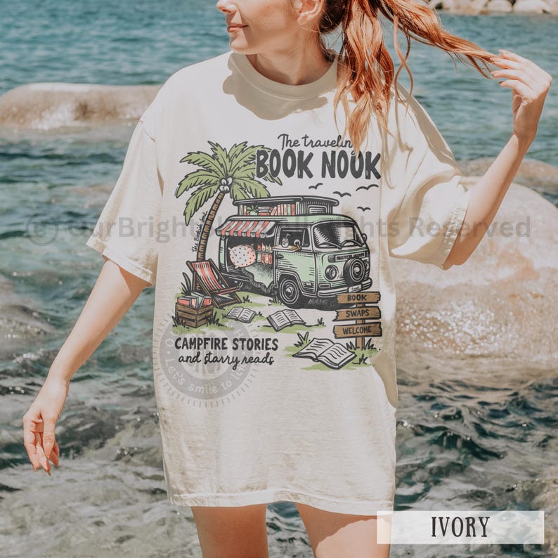 May include: A light ivory t-shirt with a graphic of a green vintage van with a sign that says "The Travelin' Book Nook". The van is parked in front of a palm tree and has books stacked inside. The graphic also includes the text "Campfire Stories and Starry Reads".