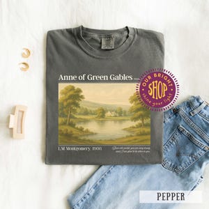 May include: Pepper-colored t-shirt featuring a landscape painting from "Anne of Green Gables" with text. The design includes a lake, trees, and a house. The text reads "Anne of Green Gables" and "L.M. Montgomery, 1908".