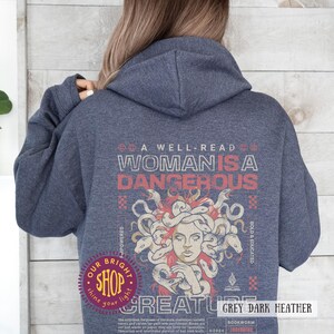 Op de afbeelding: Een donkergrijze hoodie met een afbeelding van Medusa en de tekst "A Well-Read Woman is a Dangerous Creature." Het ontwerp bevat rode en witte tekst en een shoplogo. De hoodie is gemaakt van een zacht, comfortabel materiaal.