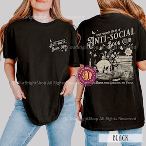 May include: Black t-shirt with the text "UNAPOLOGETICALLY ANTI-SOCIAL BOOK CLUB" on the front and back. The back graphic includes a skull, books, and a teacup. The shirt is a comfortable, casual style.