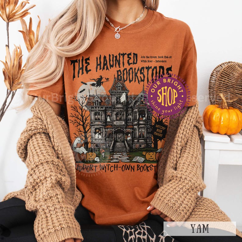 以下が含まれることがあります： 「THE HAUNTED BOOKSTORE」のテキストと、お化け屋敷のグラフィックが特徴のオレンジ色のTシャツ。 デザインには、ほうきに乗った魔女、幽霊、カボチャ、「Witchy Book Awards」の文字が含まれています。