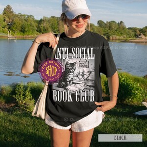 May include: Black t-shirt with the words "ANTI-SOCIAL BOOK CLUB" and a graphic of a cat sitting on a book. The cat has a "DO NOT DISTURB" sign. The shirt is a casual style.