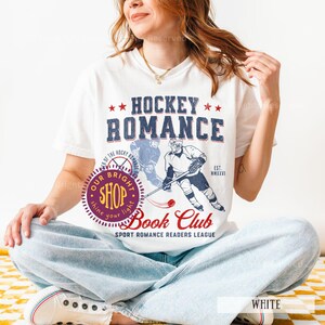 May include: White t-shirt with a Hockey Romance graphic. The design features the text "Hockey Romance" above an illustration of a hockey player. The shirt also has the text "Book Club" and "Sport Romance Readers League".