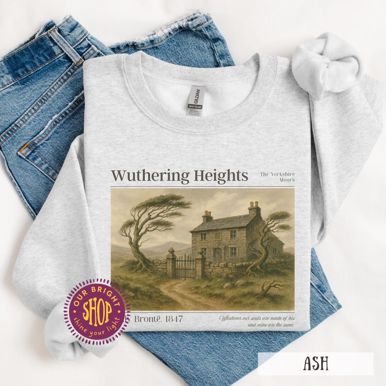 Puede incluir: Sudadera gris ceniza con el texto "Wuthering Heights" y una ilustraci&oacute;n de una casa de piedra. Tambi&eacute;n se ve el texto "Bront&euml;, 1847". La sudadera est&aacute; doblada sobre unos vaqueros azules.