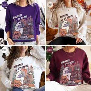 May include: Crewneck sweatshirts in purple, sand, white, and maroon feature a Halloween-themed graphic. The design includes a ghost reading a book, pumpkins, and the text "HAUNTED BY BOOKS". The sweatshirts are a cozy fall fashion item.