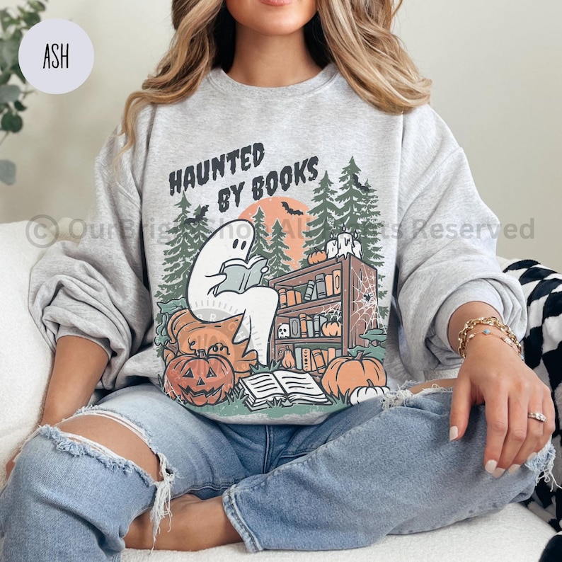 May include: Ash gray sweatshirt with a Halloween-themed graphic. The design features a ghost reading a book in front of a bookshelf, pumpkins, and the text "HAUNTED BY BOOKS". The graphic is in shades of orange, green, and black.