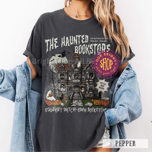 以下が含まれることがあります： 「THE HAUNTED BOOKSTORS」のテキストが入った、お化け屋敷のイラストが描かれた濃いグレーのTシャツ。デザインには魔女、カボチャ、幽霊が含まれています。シャツには「SUPPORT WITCH-OWN BOOKSTORS」とも書かれています。