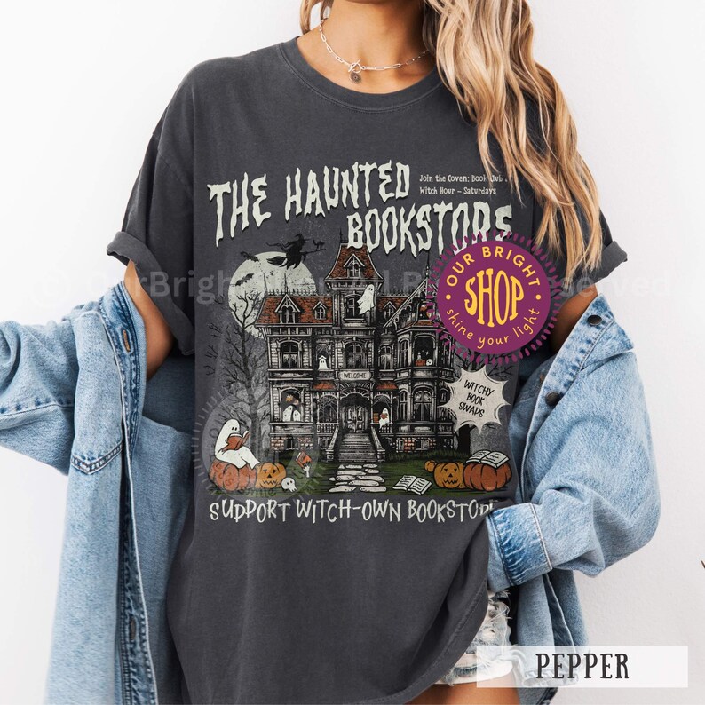 以下が含まれることがあります： 「THE HAUNTED BOOKSTORS」のテキストが入った、お化け屋敷のイラストが描かれた濃いグレーのTシャツ。デザインには魔女、カボチャ、幽霊が含まれています。シャツには「SUPPORT WITCH-OWN BOOKSTORS」とも書かれています。