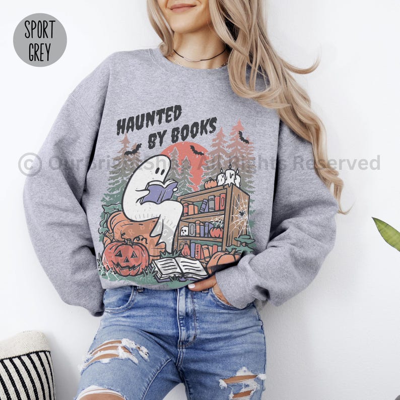 May include: Sport grey sweatshirt with a Halloween-themed graphic. The design features a ghost reading a book in front of a bookshelf, pumpkins, and the text "HAUNTED BY BOOKS". The background includes a red moon and trees.