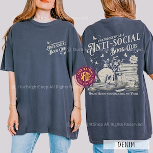 May include: Denim blue t-shirt with the text "UNAPOLOGETICALLY ANTI-SOCIAL BOOK CLUB" on the front and back. The back graphic features a skull, books, and the phrase "MAKING FRIENDS WITH CHARACTERS, NOT PEOPLE."