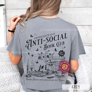 May include: A grey t-shirt with a graphic design that reads "UNAPOLOGETICALLY ANTI-SOCIAL BOOK CLUB". The design includes a skull, books, butterflies, and the text "MAKING FRIENDS WITH CHARACTERS, NOT PEOPLE".