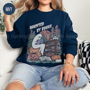 May include: Navy blue sweatshirt with a Halloween-themed graphic. The design features a ghost reading a book in front of a bookshelf, pumpkins, and the text "HAUNTED BY BOOKS". The sweatshirt has a crew neck and long sleeves.