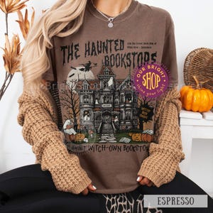 以下が含まれることがあります： エスプレッソ色のTシャツ。お化け、カボチャ、ほうきに乗った魔女が描かれた、お化け屋敷の書店をテーマにした不気味なイラスト。テキストには「The Haunted Bookstores」と「Witchy Book Snaps」と書かれています。