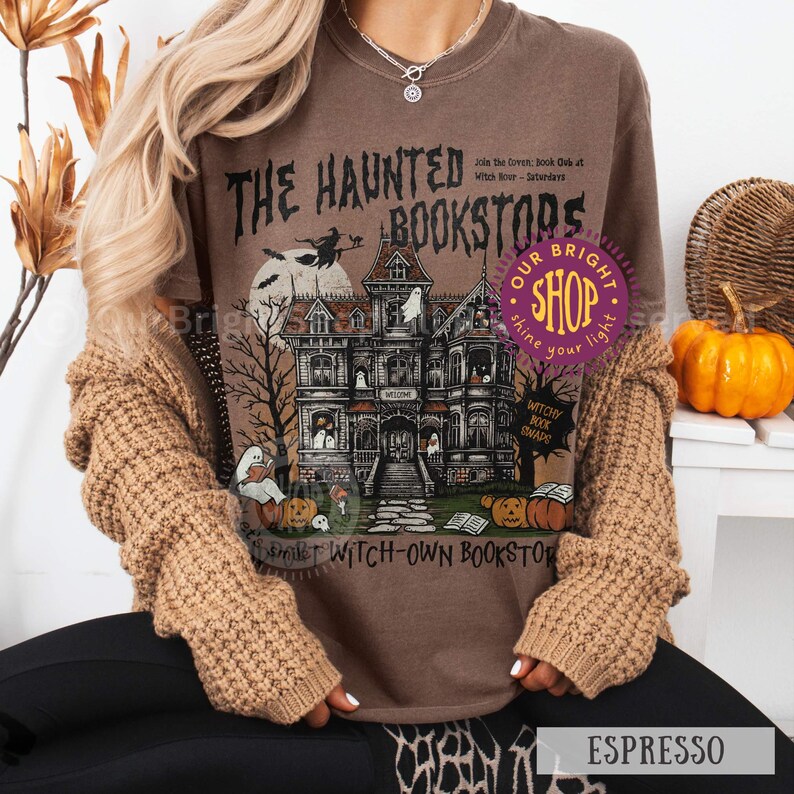 以下が含まれることがあります： エスプレッソ色のTシャツ。お化け、カボチャ、ほうきに乗った魔女が描かれた、お化け屋敷の書店をテーマにした不気味なイラスト。テキストには「The Haunted Bookstores」と「Witchy Book Snaps」と書かれています。