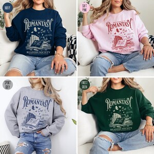 May include: A close-up of four sweatshirts in different colors: navy blue, soft pink, sport grey, and forest green. Each sweatshirt features a graphic design with the text "Proud Member of the Romantasy Readers Society" and an illustration of a stack of books, butterflies, and stars.