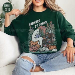 May include: Forest green sweatshirt with the text "HAUNTED BY BOOKS" and a graphic of a ghost reading a book, surrounded by pumpkins, a bookshelf, and trees. The sweatshirt is a crew neck style.