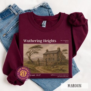 Puede incluir: Sudadera granate con el texto "Wuthering Heights" y una imagen de una casa de piedra y &aacute;rboles. El texto "Bront&euml;. 1817" est&aacute; debajo. La sudadera est&aacute; doblada sobre unos vaqueros azules.