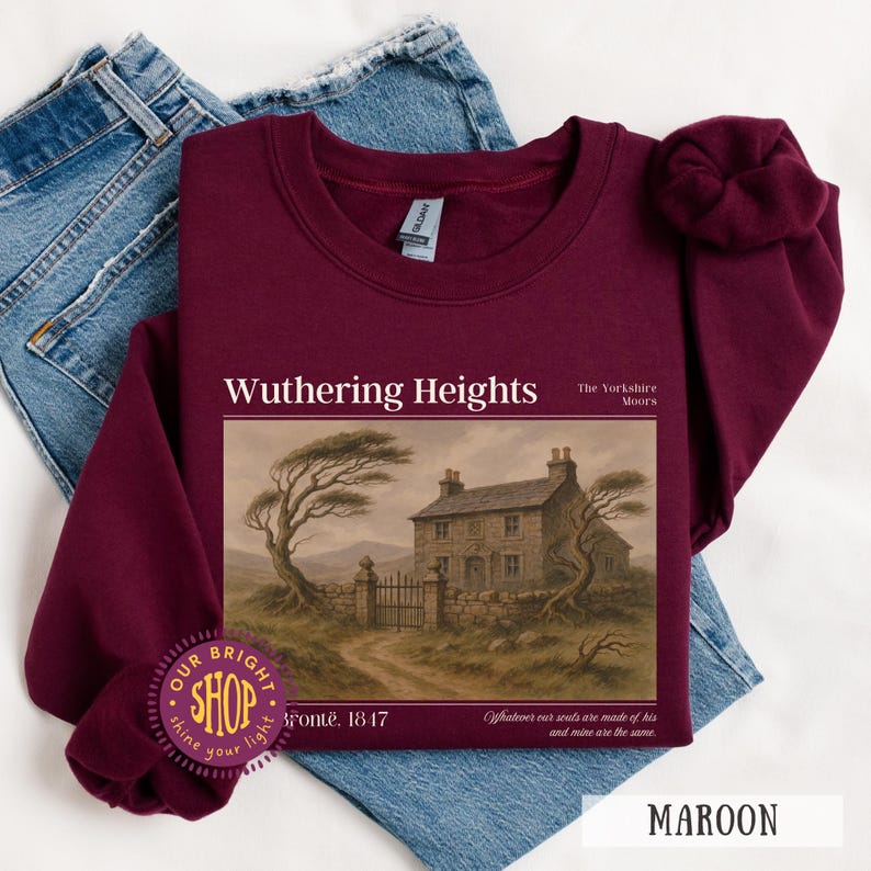 Puede incluir: Sudadera granate con el texto "Wuthering Heights" y una imagen de una casa de piedra y &aacute;rboles. El texto "Bront&euml;. 1817" est&aacute; debajo. La sudadera est&aacute; doblada sobre unos vaqueros azules.