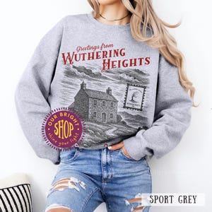 May include: Sport grey sweatshirt featuring a graphic of a house and the text "Greetings from Wuthering Heights." The design includes a postage stamp graphic and the shop's logo. The sweatshirt is paired with blue jeans.