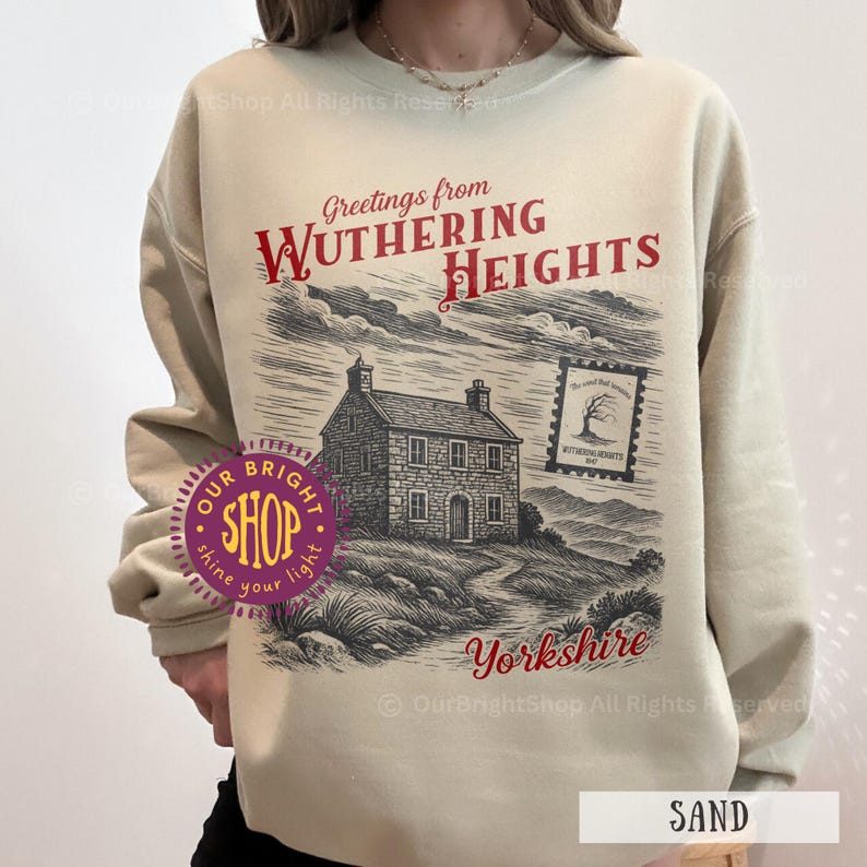 May include: Sand-colored sweatshirt featuring a graphic of a house on a hill with the text "Greetings from Wuthering Heights" in red. The word "Yorkshire" is also printed in red. A stamp graphic is also visible.