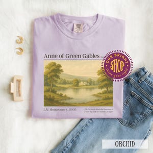 May include: Orchid-colored t-shirt featuring a landscape painting from "Anne of Green Gables" by L.M. Montgomery. The shirt includes the text "Anne of Green Gables" and a quote. A pair of gold hoop earrings, a hair clip, and jeans are also in the image.