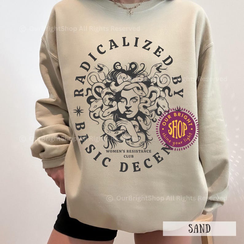 Peut inclure: Sweat-shirt couleur sable avec un motif circulaire repr&eacute;sentant M&eacute;duse et le texte "RADICALIZED BY BASIC DECENCY". Le motif comprend le texte "WOMEN'S RESISTANCE CLUB" et un logo de magasin violet. Le mot "SAND" est imprim&eacute; en bas.