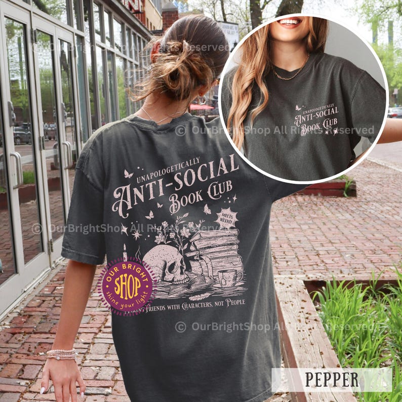 May include: Pepper-colored t-shirt with a graphic design featuring a skull, books, and the text "Unapologetically Anti-Social Book Club." The design includes butterflies and the phrase "Friends with Characters, Not People."