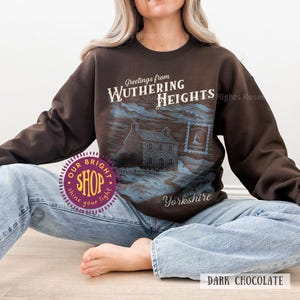 May include: Dark chocolate-colored sweatshirt featuring a graphic of a house and the text "Greetings from Wuthering Heights Yorkshire." The design includes a postage stamp illustration. The sweatshirt is a crew neck style.