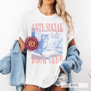May include: White t-shirt with the text "ANTI-SOCIAL BOOK CLUB" in pink and blue. The graphic features a cat sitting by a window with a "DO NOT DISTURB" sign. The shirt also has a shop logo.