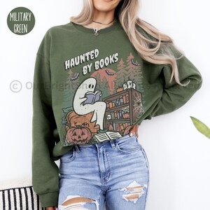 May include: Military green sweatshirt with the text "HAUNTED BY BOOKS" and a graphic of a ghost reading a book. The design includes pumpkins, bats, and a bookshelf, perfect for Halloween or book lovers.