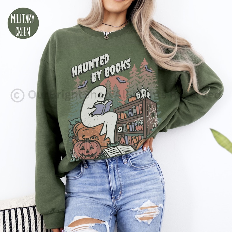 May include: Military green sweatshirt with the text "HAUNTED BY BOOKS" and a graphic of a ghost reading a book. The design includes pumpkins, bats, and a bookshelf, perfect for Halloween or book lovers.