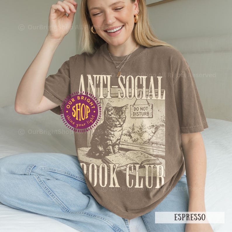 May include: Espresso-colored t-shirt with the text "ANTI-SOCIAL BOOK CLUB" and a graphic of a cat sitting by a window with a book. The shirt also has the words "DO NOT DISTURB" on a sign.