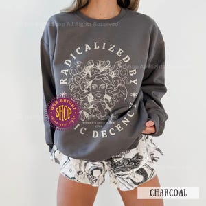 Peut inclure: Sweat-shirt gris anthracite avec un motif blanc de M&eacute;duse et le texte "RADICALIZED BY IC DECENCY WOMEN'S RESISTANCE CLUB". Le mod&egrave;le porte un short blanc avec un imprim&eacute; graphique noir. Le mot "CHARCOAL" est visible.