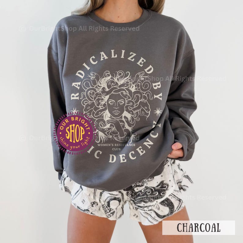 Peut inclure: Sweat-shirt gris anthracite avec un motif blanc de M&eacute;duse et le texte "RADICALIZED BY IC DECENCY WOMEN'S RESISTANCE CLUB". Le mod&egrave;le porte un short blanc avec un imprim&eacute; graphique noir. Le mot "CHARCOAL" est visible.