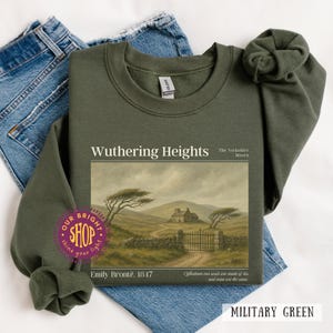May include: Military green sweatshirt featuring the title "Wuthering Heights" with an illustration of a landscape. The text "The Yorkshire Moors", "Emily Brontë. 1847", and a quote are also visible. The sweatshirt is displayed on top of blue jeans.