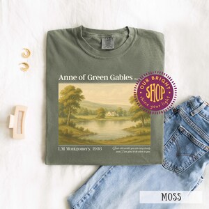 May include: Moss green t-shirt featuring a landscape painting from "Anne of Green Gables" by L.M. Montgomery. The shirt includes the text "Anne of Green Gables" and "L.M. Montgomery, 1908". The shirt is displayed with blue jeans and accessories.