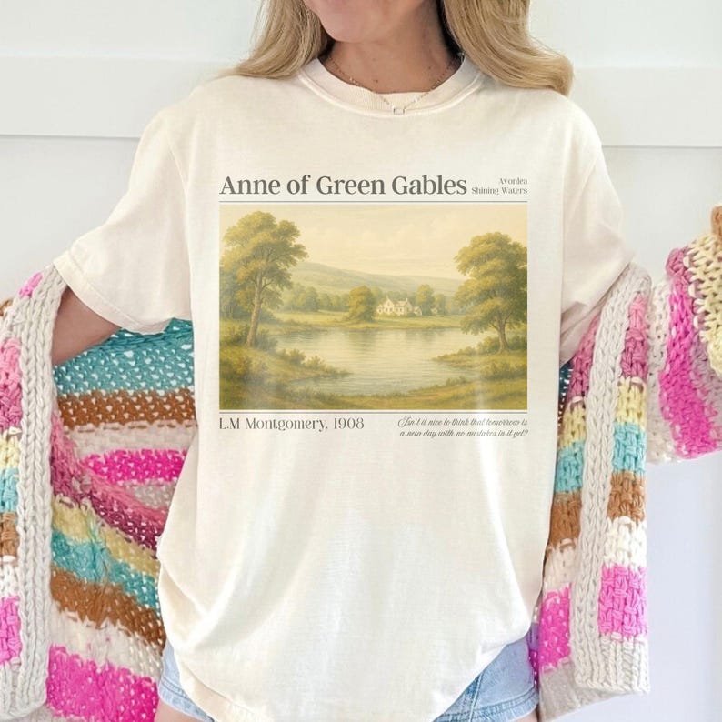 May include: Cream-colored t-shirt featuring a landscape painting from "Anne of Green Gables" by L.M. Montgomery. The artwork depicts a serene lake scene with trees and a house. The text "Anne of Green Gables" and the author's name are printed above the image.