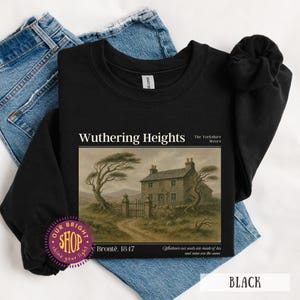 Puede incluir: Sudadera negra con el t&iacute;tulo "Wuthering Heights" y una ilustraci&oacute;n de una casa y un paisaje. Tambi&eacute;n se ve el texto "The Yorkshire Moors" y "Bront&euml;. 1847". La sudadera se muestra sobre unos vaqueros azules.