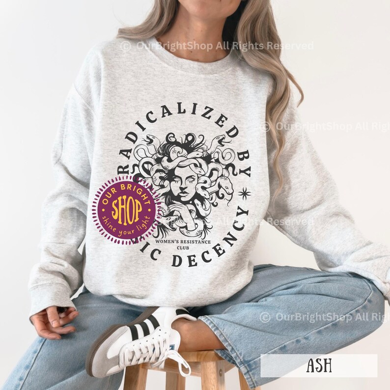 Peut inclure: Sweat-shirt gris clair avec un motif de M&eacute;duse entour&eacute; du texte "RADICALIZED BY IC DECENCY" et "WOMEN'S RESISTANCE CLUB". Un logo de boutique circulaire est &eacute;galement visible.