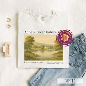 May include: White t-shirt featuring the text "Anne of Green Gables" above a landscape painting. The shirt includes the text "L.M. Montgomery, 1908" and a quote. A purple shop logo is visible. The shirt is paired with blue jeans.