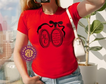 May include: A red t-shirt featuring a black graphic of a locket with the letters "HC" inside. The locket is adorned with a bow and ribbon. The text "Whatever our souls are made of, his and mine are the same" is also printed on the shirt.